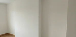 Corner detail showing a seamless plasterboard media wall merging perfectly with an existing room wall, featuring a monolithic skim-coat finish with no visible joints.